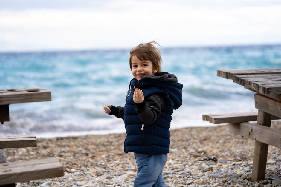 Young boy with brown hair in winter clothing smiling at a pebble beach in Aydın, Türkiye.