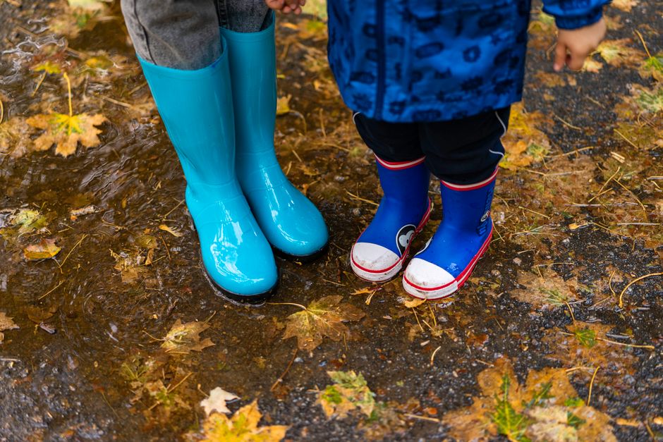 Two children wearing bright rubber boots splash in autumn puddles covered with fallen leav