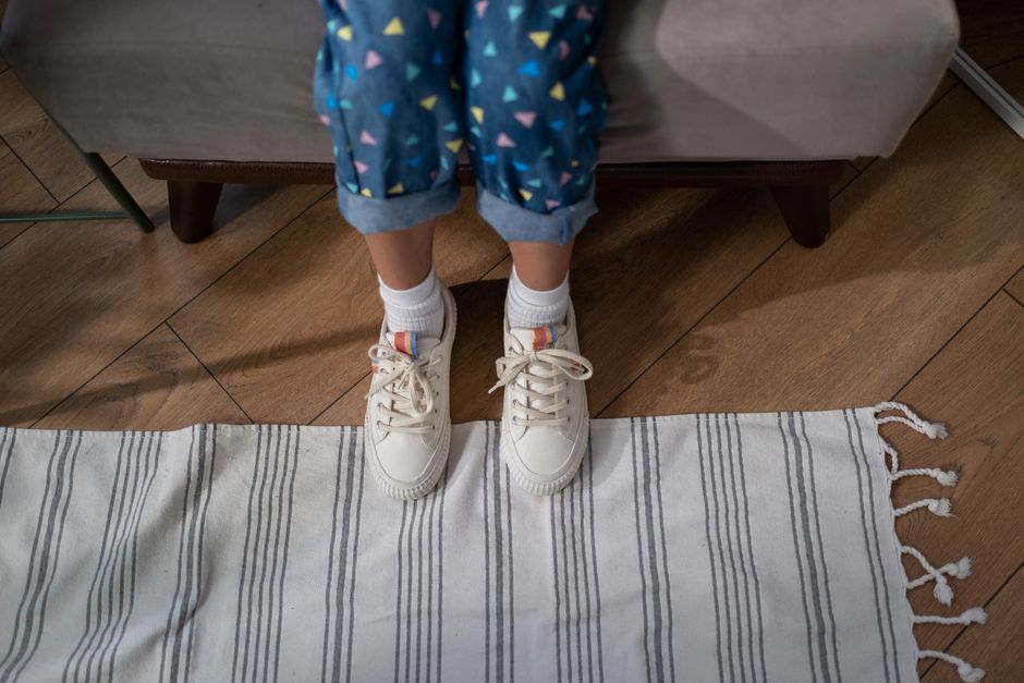 Child standing on a rug wearing patterned pants and sneakers in a cozy indoor setting.