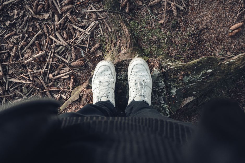 A top view of a person standing in white sneakers on a forest ground with pinecones.