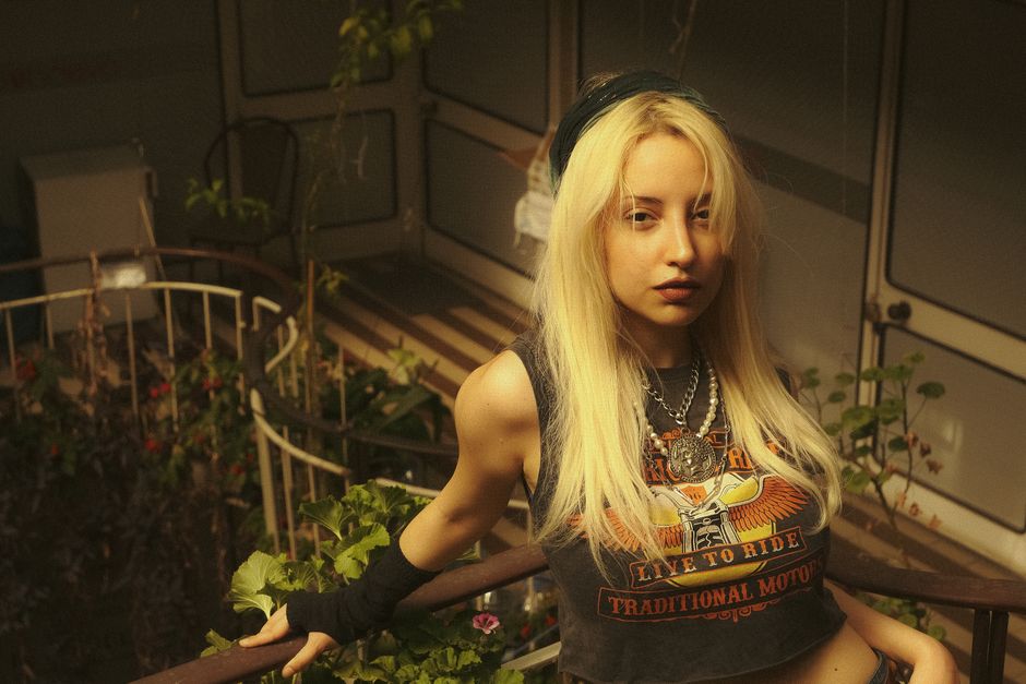 Blonde woman posing in an indoor garden wearing a biker-themed top, exuding a modern and edgy vibe.